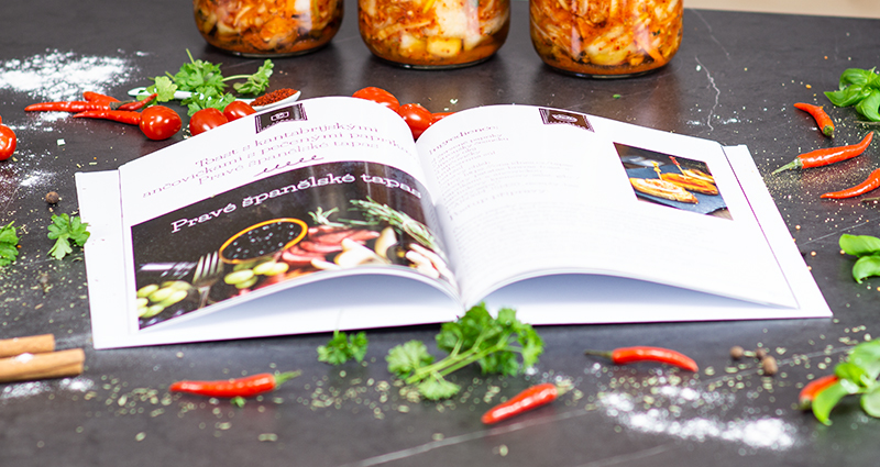 Detailný záber na fotoknihu s receptami, v pozadí poháre so zaváranou zeleninou, okolo chilli papričky, cherry paradajky a korenie.