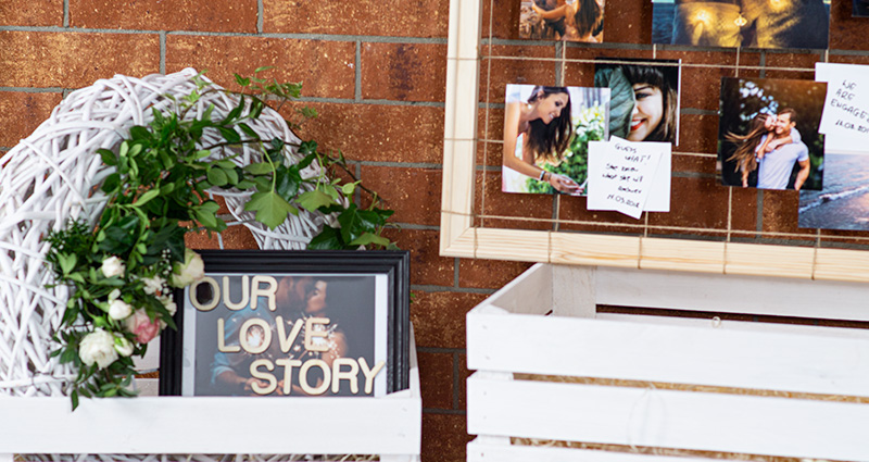 Zoom sobre la inscripción Our Love Story en el marco negro decorado con flores, en una caja de madera blanca. Al lado fragmento de un marco de bricolaje de madera con fotos de los novios de varias etapas de su vida, y junto con unos comentarios. En el fondo una pared de ladrillos. 