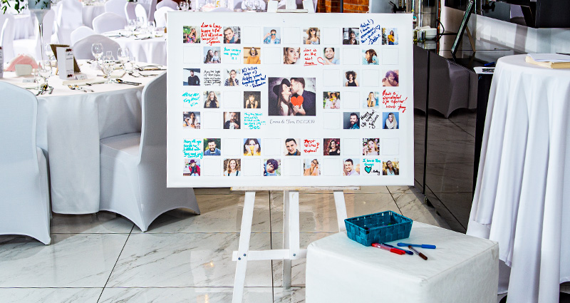 Lienzo horizontal con una foto de los novios de la sesión de compromiso en el medio y fotos de invitados alrededor, y los lugares para sus firmas/notas. Lienzo sobre un caballete blanco, con una pequeña mesa frente a él, y una canasta con marcadores coloridos. En el fondo hay mesas redondas en el salón de bodas. 