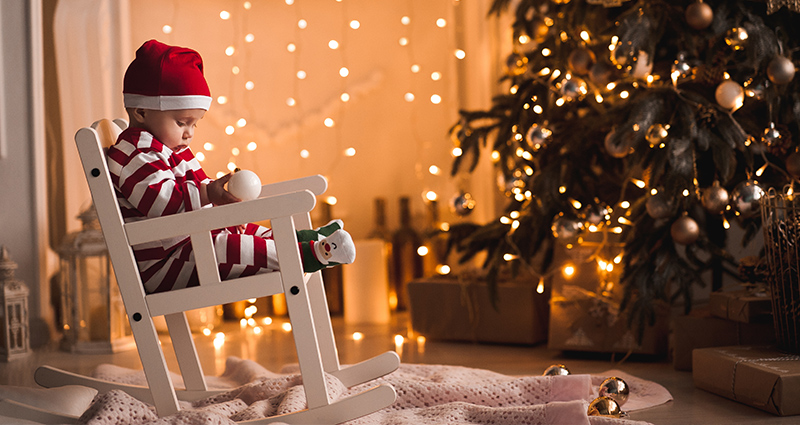 Een jongetje in kerstpijama en met de muts van de Kerstman op dat ligt te slapen in een schommelstoel naast de kerstboom