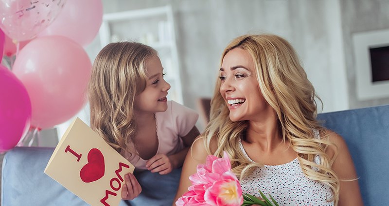 Mutter sitzt auf einem Sofa und liest eine aufgeschlagene Karte mit „ I <3 MOM” von ihrer Tochter. Tochter lehnt sich hinter der Couch an. Links ein Bündel rosa Ballons. 