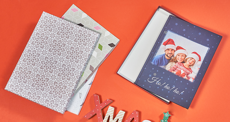 El fotolibro con la tapa azul en la caja que presenta la familia sonriente con los gorros de Papá Noel. Al lado de eso están puestas tres cajas para los fotolibros, más abajo pone la inscripción de madera XMAS. Todos los productos sobre fondo rojo.