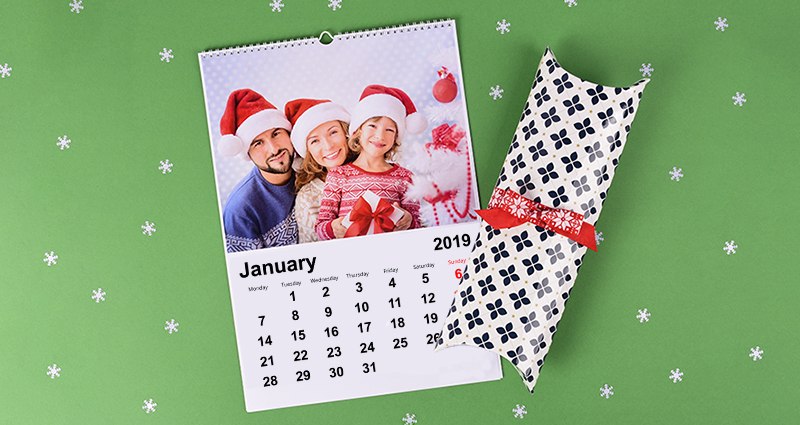 A photo calendar with a photo of a smiling family wearing Santa Claus hats; a decorative gift wrap box for a photo calendar tied with a red tow next to it. The products are lying on the green background with white stars all around.