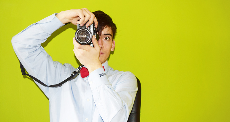 Una foto de un hombre en una camiseta azul y con una pajarita roja tomando una foto con una cámara analógica, una pared de color limón en el fondo.