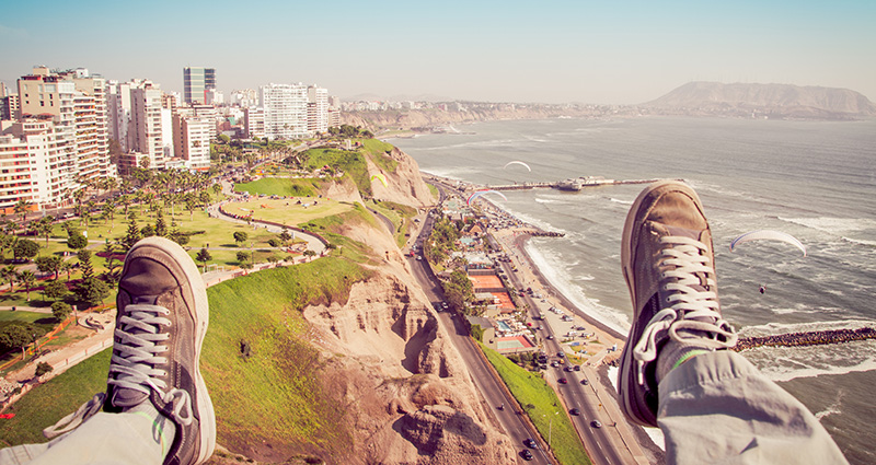 A picture of a modern city by the coast, legs of a man sitting on the edge in the foreground