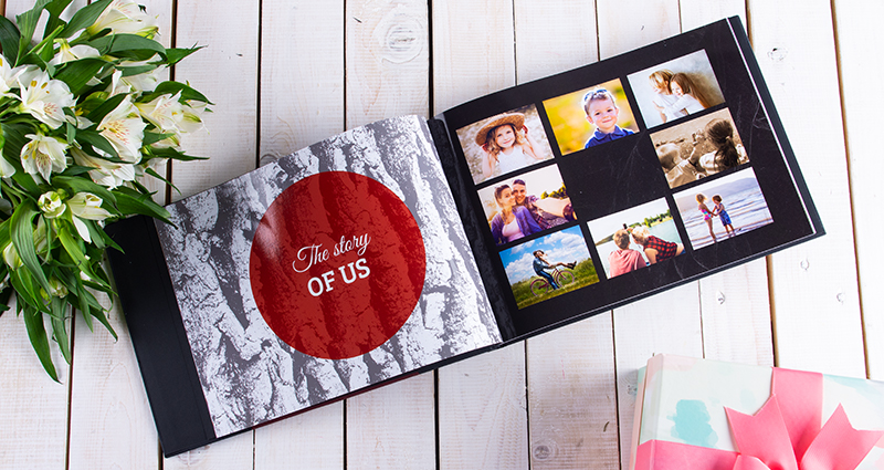 A picture of an open photo book with photos of a little girl and boy – a gift on the right side and white flowers bouquet on the left side of it.