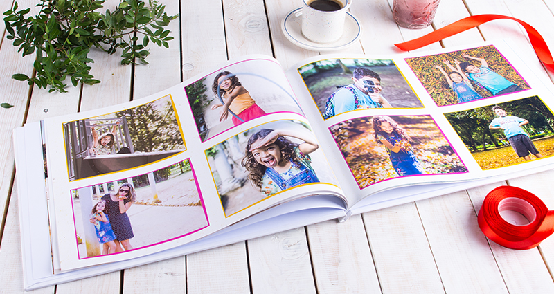 A picture of an open photo book with the photos of a little girl and boy – red ribbon, cup of coffeee, a candle and green branch next to it.