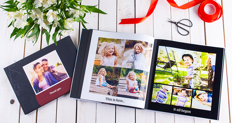 Une photo de deux livres photo - un album fermé avec la photo de couple amoureux sur la couverture et le texte „Thank you”, à côté un livre photo ouvert (à gauche les photos d'enfance de fille et à droite - de garçon). Au-dessus de l’album ouvert un rouban rouge et des ciseaux, dans le coin supérieur gauche un bouquet des fleurs blanches.