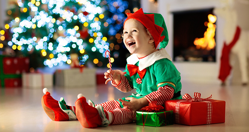 A preschooler in an elf suit next to presents and a Christmas tree