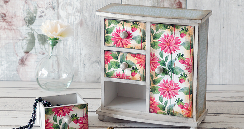 A vintage dresser with the front surface covered in pink flowers. Nearby a dresser drawer and chaplets hanging out of it. In the background on the left side white flowers in a vase.