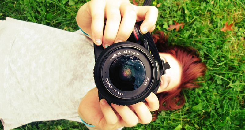 A woman in a bright blouse lying on grass and keeping her hands in the air in which she’s holding an analogue camera. 