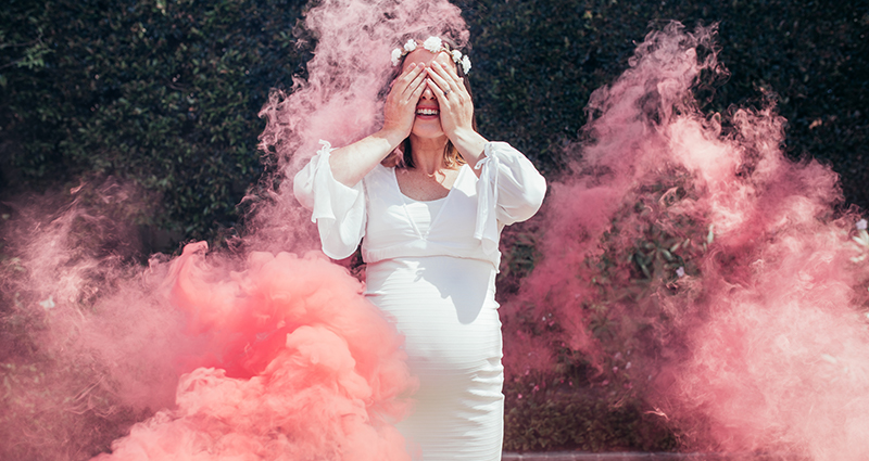 Eine junge schwangere Frau in einem engen weißen Kleid während eines Schwangerschaft-Fotoshootings mit farbigem Pulver