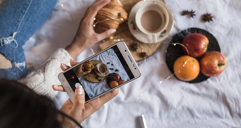 Detailný záber na ruku ženy, ktorá so smartphoneom fotografuje kávu, croissant a ovocie ležiace na posteli.