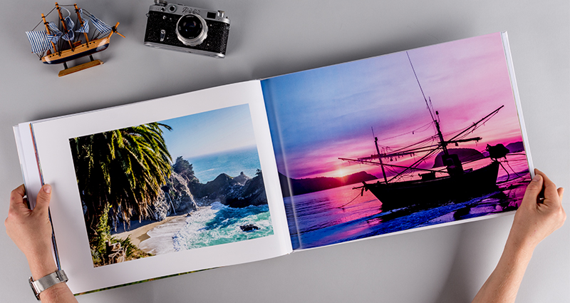 An open travel photo book A3 lying next to a ship statuette and a camera, focus on the hands of the person looking at it.