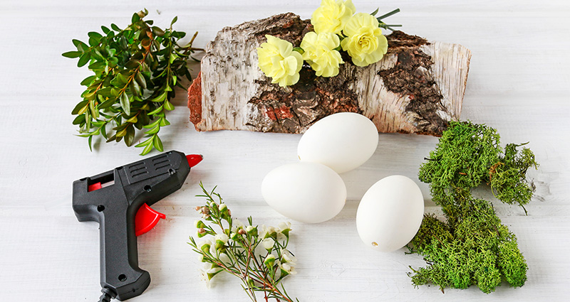 Une bûche de bouleau,des branches de buis, une  mousse, des œufs  et un pistolet avec de la colle à chaud sur une table claire.