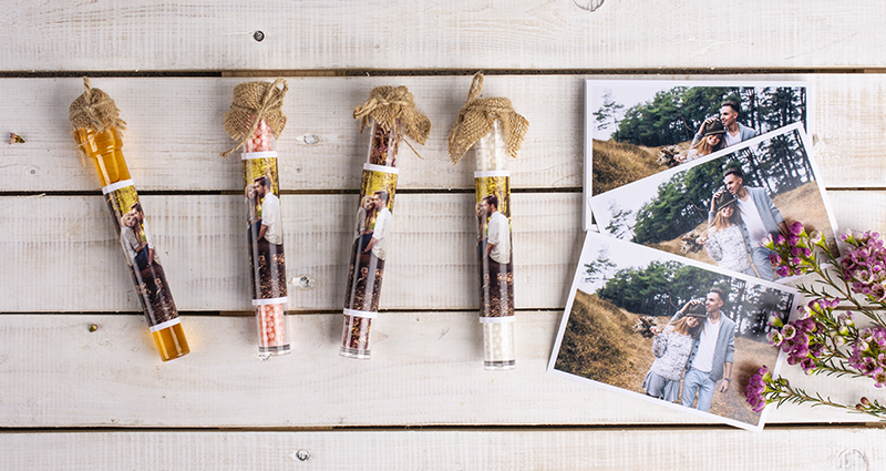 Coffee and sweets in a colourful containers decorated with jute and prints, 3 photos and tiny pink flowers next to it . A composition lays on white wooden planks.