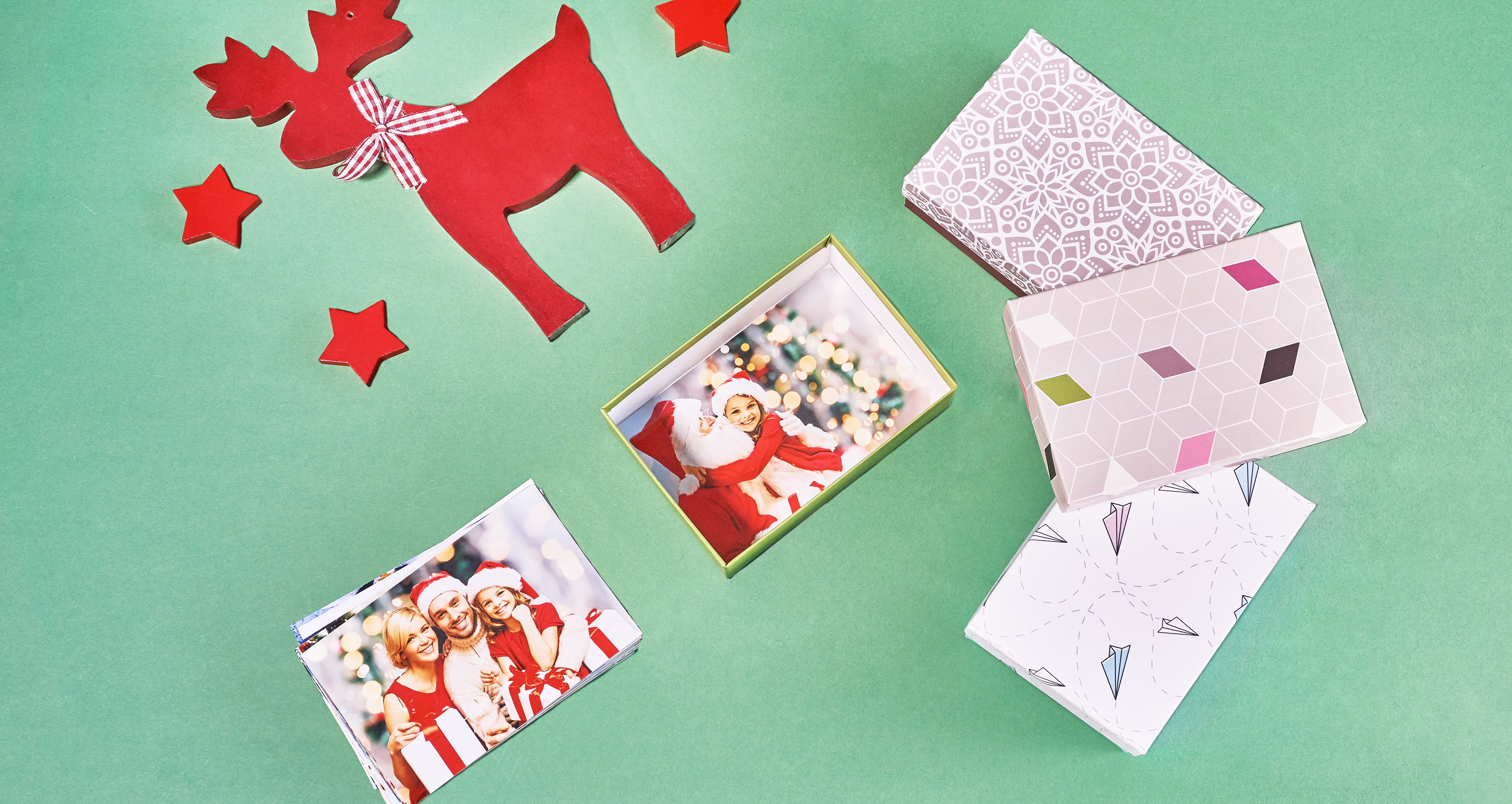 Les tirages classiques dans une boîte cadeau, à côté un tas de photos. À droite 3 emballages décoratifs pour les tirages, au dessus une renne rouge en bois et les étoiles. Tous les produits sur un fond vert.
