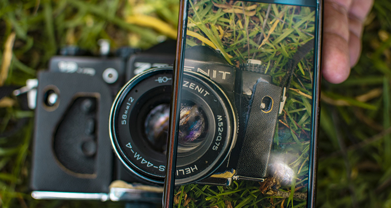 Detailní záběr na smartphone, na kterých displeji je vidět starý fotoaparát ležící na trávě.