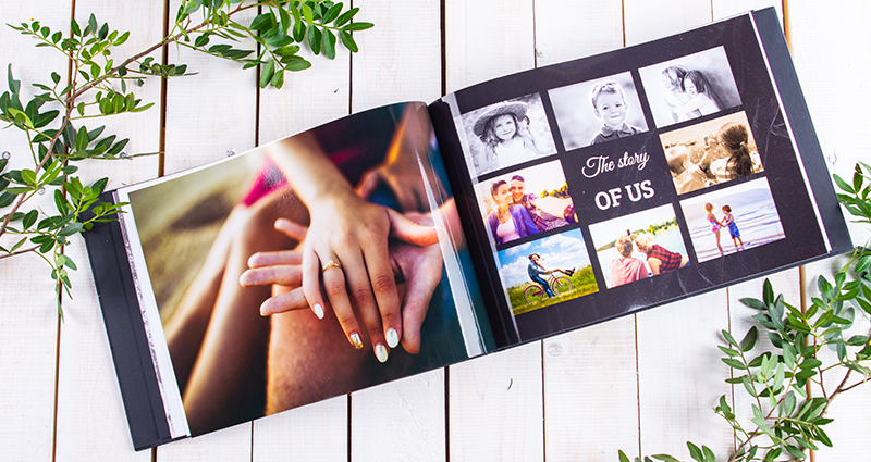 Close up on an open photo book – photo of couple's hands (engagement ring on woman's ring finger) on the left side, a collage on the other page. A photo book on a bright wooden background decorated with green branches. 