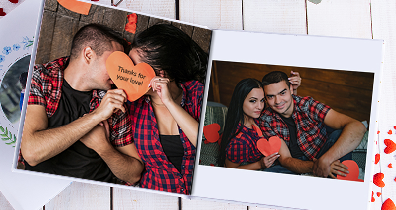 Close up on two Luxury Albums. Pictures of a couple holding paper hearts with captions in an open album. Red decorative hearts and a branch next to the albums.