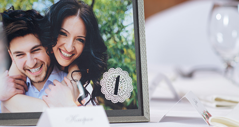 Close-up on a photo of the bride and groom, from the wedding photo session, in a black-silver frame with number 1 put on a wedding table. Tableware and name cards around.