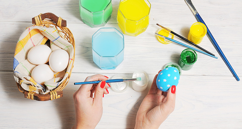 Close-up on a woman’s hands painting an Easter egg. Eggs in a basket, colourful paints and brushes laying around.