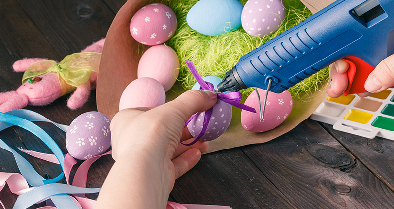 Zoom sobre la mano de una persona que está decorando unos huevos de Pascua, al lado unas pinturas y unas cintas de tela de colores.