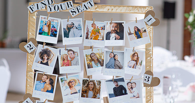 Close-up on the upper part of the handmade photo frame with photos of the wedding guests near specific table numbers. “Find your seat” caption at the top. Ballroom table in the background.