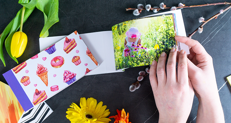 Detailný záber na ruky ženy prezerajúcej sharebook s jarnými fotografiami dieťaťa. Vedľa Sharebooky v rozných obaloch. Okolo tulipány, gerbery a bahniatka.