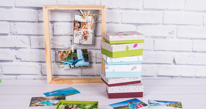 Collection of cardboard boxes for photo prints in two formats organized one on another, next to them there is a frame with photos hanged on a wooden clips, spread prints in the front, brick wall in the background.