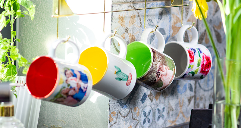 Les mugs colorés de printemps suspendus à des crochets au dessus de plan de travail, à côté une vase avec des fleurs de printemps.