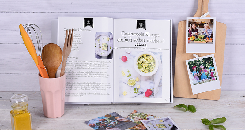 Al centro un libro di ricette, accanto stampe retrò su un tagliere, una  tazza con degli utensili da cucina, altre foto e delle mele.