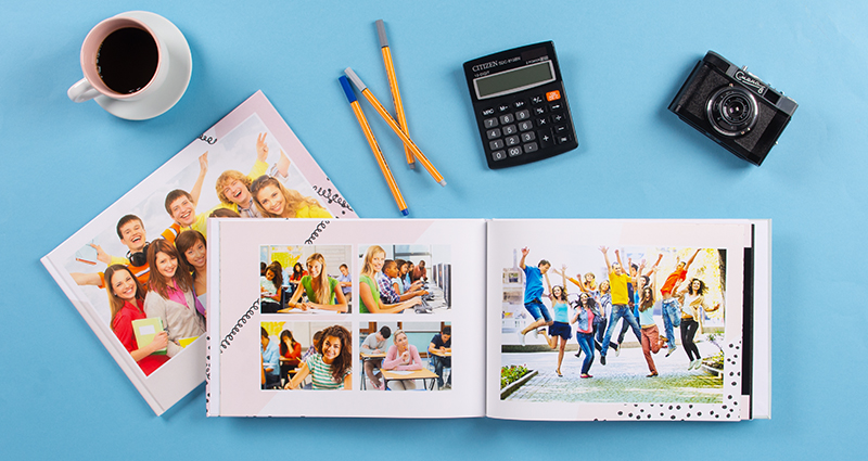 Dve školské fotoknihy (jedna otvorená, druha uzavretá) na modrom pozadí. Nad nimi šálka kávy, farebné pera, kalkulačka a analógový fotoaparát.