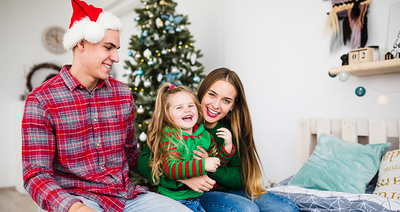 Una familia de tres personas en vestidos navideños sobre la cama, un árbol navideño en el fondo.