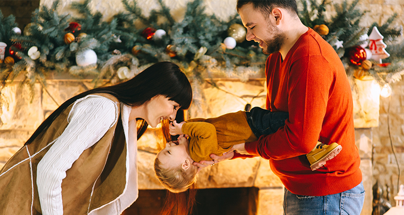 Family of three joking in front of the fireplace decorated with Christmas twigs and balls.