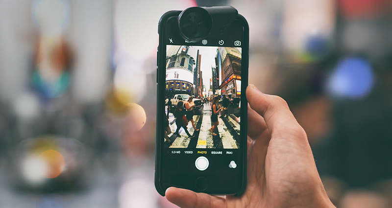 Focus on a hand holding a smartphone with a portable lens, people walking through the street on the screen; a blurred background