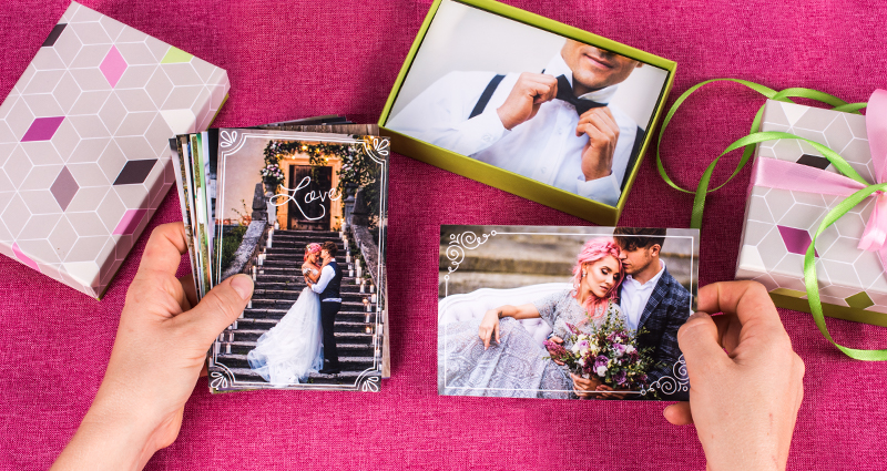 Focus on hands holding wedding photo prints, next to them one open box for photo prints, second closed with colourful ribbons, pink table cloth in the background.