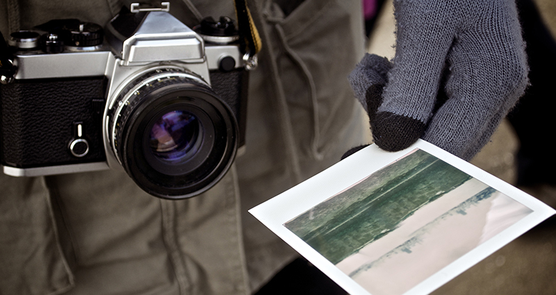 Focus on the body of a man holding a camera with a strap; there is a retro print in his gloved hand