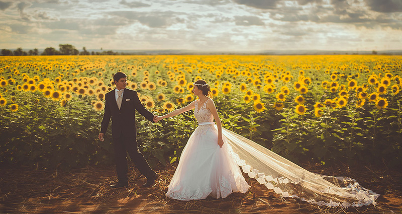 Une photo de Jeunes Mariés qui se promènent à côté de champ de tournesols, au fond le ciel nuageux.