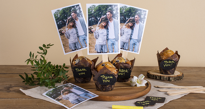 Les photos rectangulaires sur des bâtons des glaces et enfoncées dans les muffins avec le texte ”Thank you”. Les muffins sur un plateau en bois sur une nappe claire et sur une table foncée, à côté un tirage, une brindille verte, une craie jaune, des bâtons des glaces et des cartes avec des remerciements.