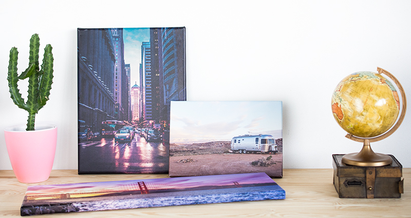 Fotolienzos de vacaciones sobre una mesa, un lienzo panorámico dispuesto sobre la mesa, dos otros contra la pared; un cactus en una maceta rosada a la izquiera, un globo terráqueo sobre una caja retro a la derecha. 