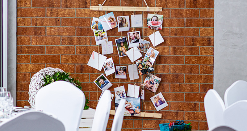 Instafotos de los invitados a la boda adjuntas a una cuerda de yute en listones de madera colgados en una pared de ladrillos. En primer plano las mesas de boda decoradas en blanco. 