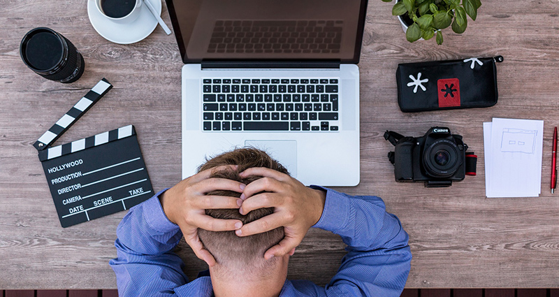 Man sitting in front of a computer and holding his head; camera, lens and other things laying next to him