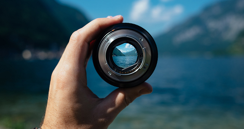 Man’s hand holding a lens in the centre, lake and mountains in the background