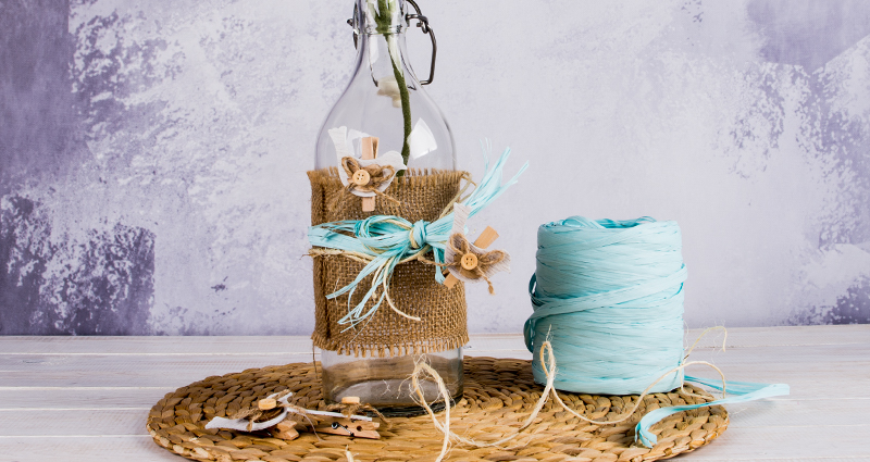 Meadow flowers in a bottle decorated with jute, raphia and wooden birds. Along side a spool of raphia and other additions.