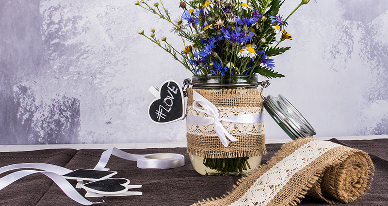 Fleurs sauvages dans un pot décoré avec  un ruban de jute et d’un clips, à côté d'un rouleau de ruban de jute et d’ autres décorations.