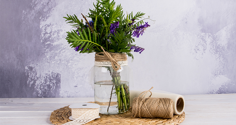 Feldblumen im Glas, das mit einer kleinen Rüsche, Spitze und Jutekordel geschmückt ist. Daneben liegen die Materialien.