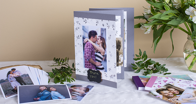 Mini scapbook with the photos of a couple and a 'Thank you' caption on the first page. Prints, sharebooks, green brunches and a white flowers bouquet in a vase next to it. A composition on a white tablecloth.