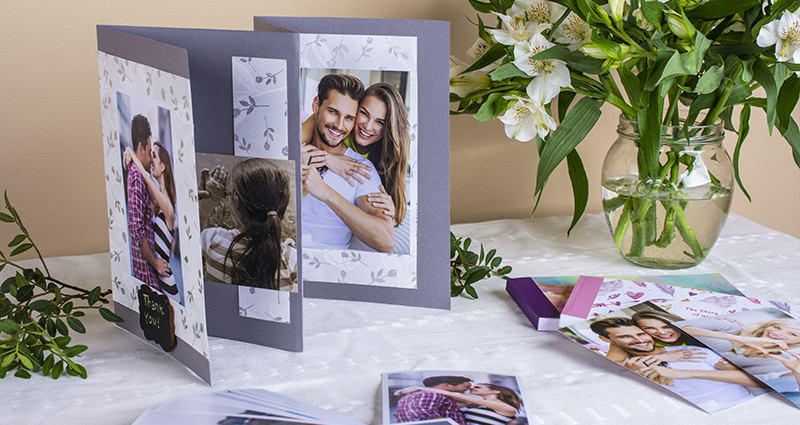 Mini-album scrapbooking avec une photo de couple amoureux et avec le texte „Thank you” sur la première page. Autour des tirages, des sharebook, des brindilles vertes et un bouquet des fleurs blanches dans una vase. La composition sur une nappe blanche.