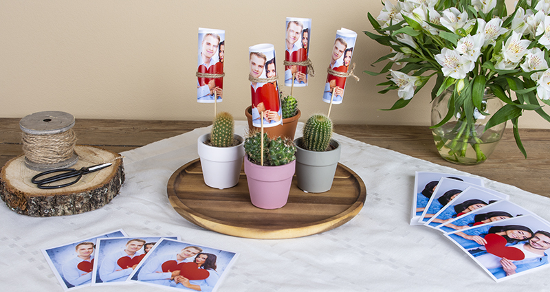 Mini cactuses in colourful pots with rolled up prints attached to skewers on a wooden tray, insta photos of a couple placed on both sides, there is white flowers bouquet in the vase, jute string and scissors. A composition on a light cloth and a dark table. 